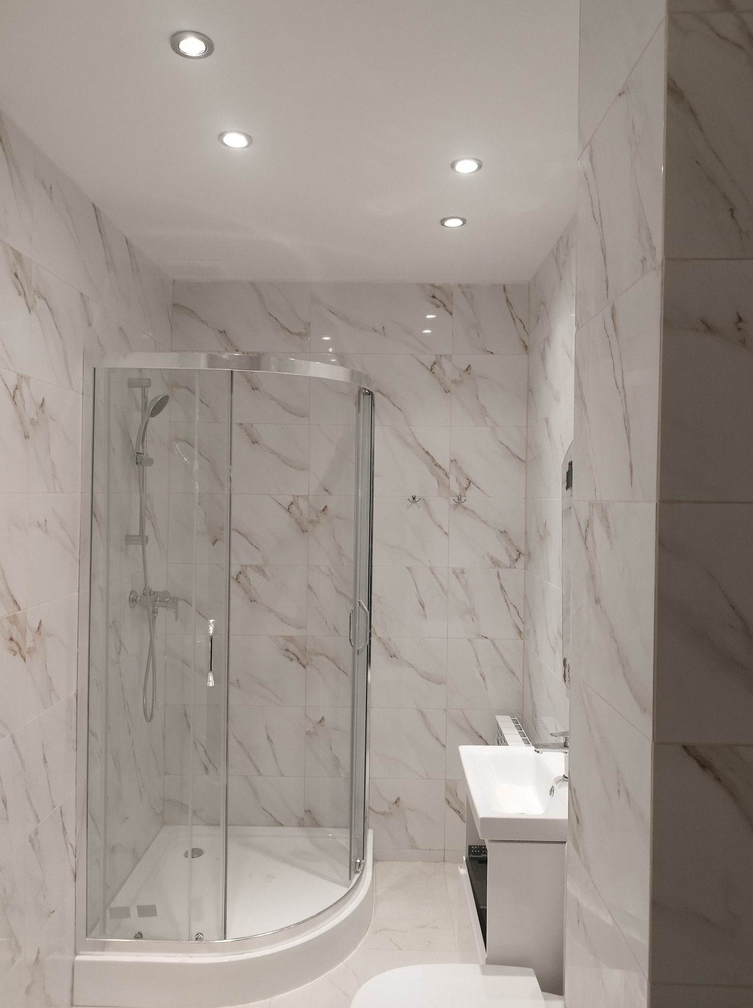 Nowoczesna łazienka z kabiną prysznicową narożną, wykończona białymi płytkami imitującymi marmur z delikatnymi brązowymi żyłami, oświetlona punktowymi lampami sufitowymi.