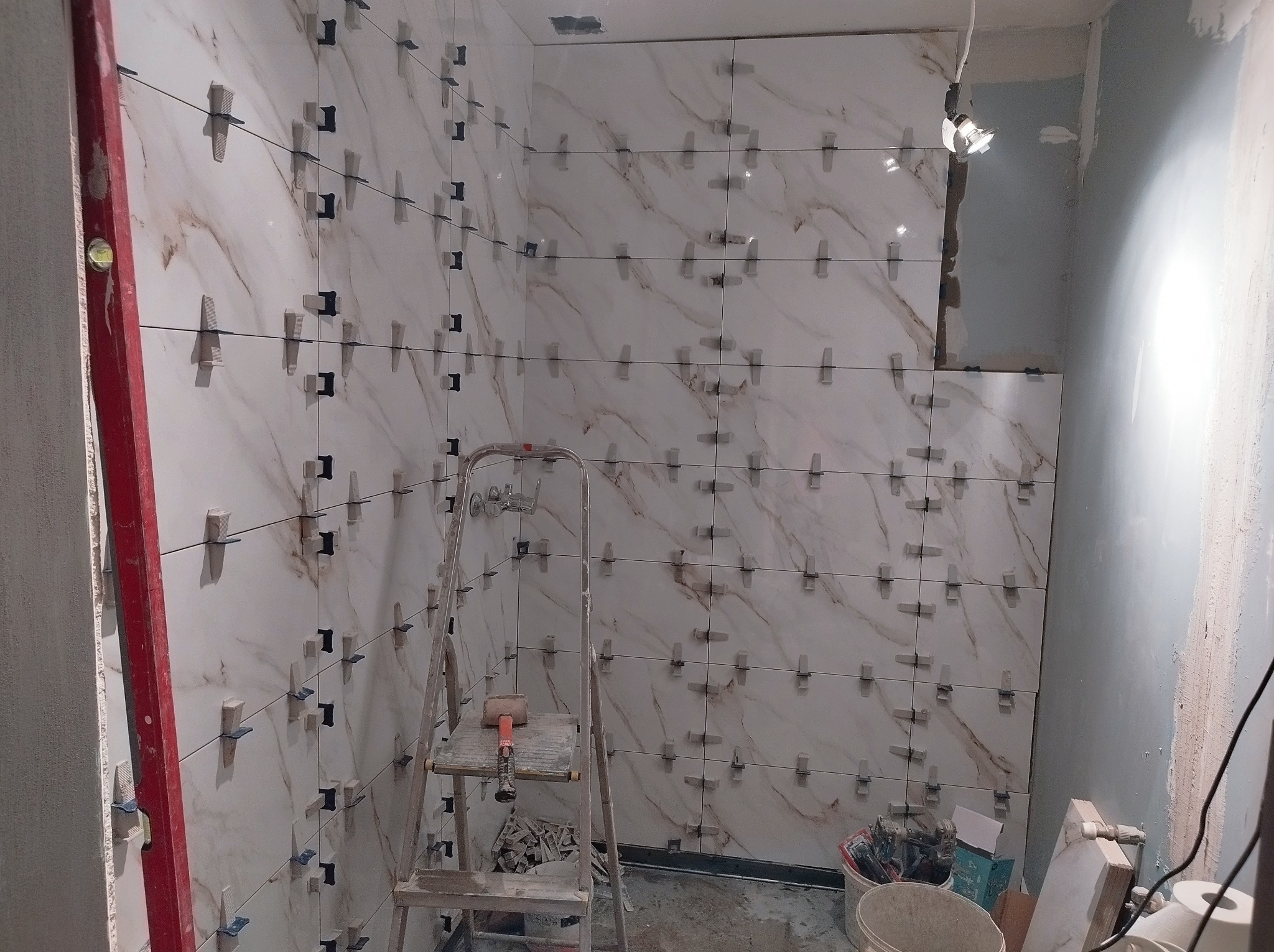 Ściana łazienki w trakcie układania marmurowych płytek, widoczne krzyżyki dystansowe, poziomica i drabina, fragment niezakończonej ściany z szarym tynkiem.