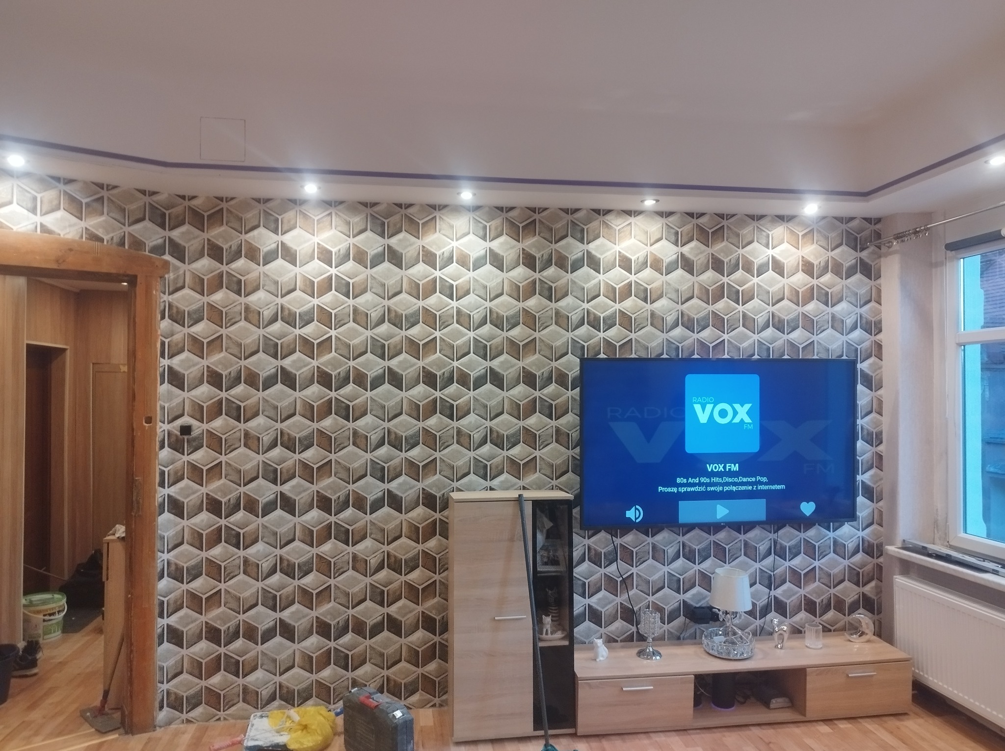 Salon po remoncie z dekoracyjną ścianą w geometryczne wzory 3D, telewizor z włączonym radiem, drewniane meble i widoczne narzędzia malarskie w rogu pomieszczenia.