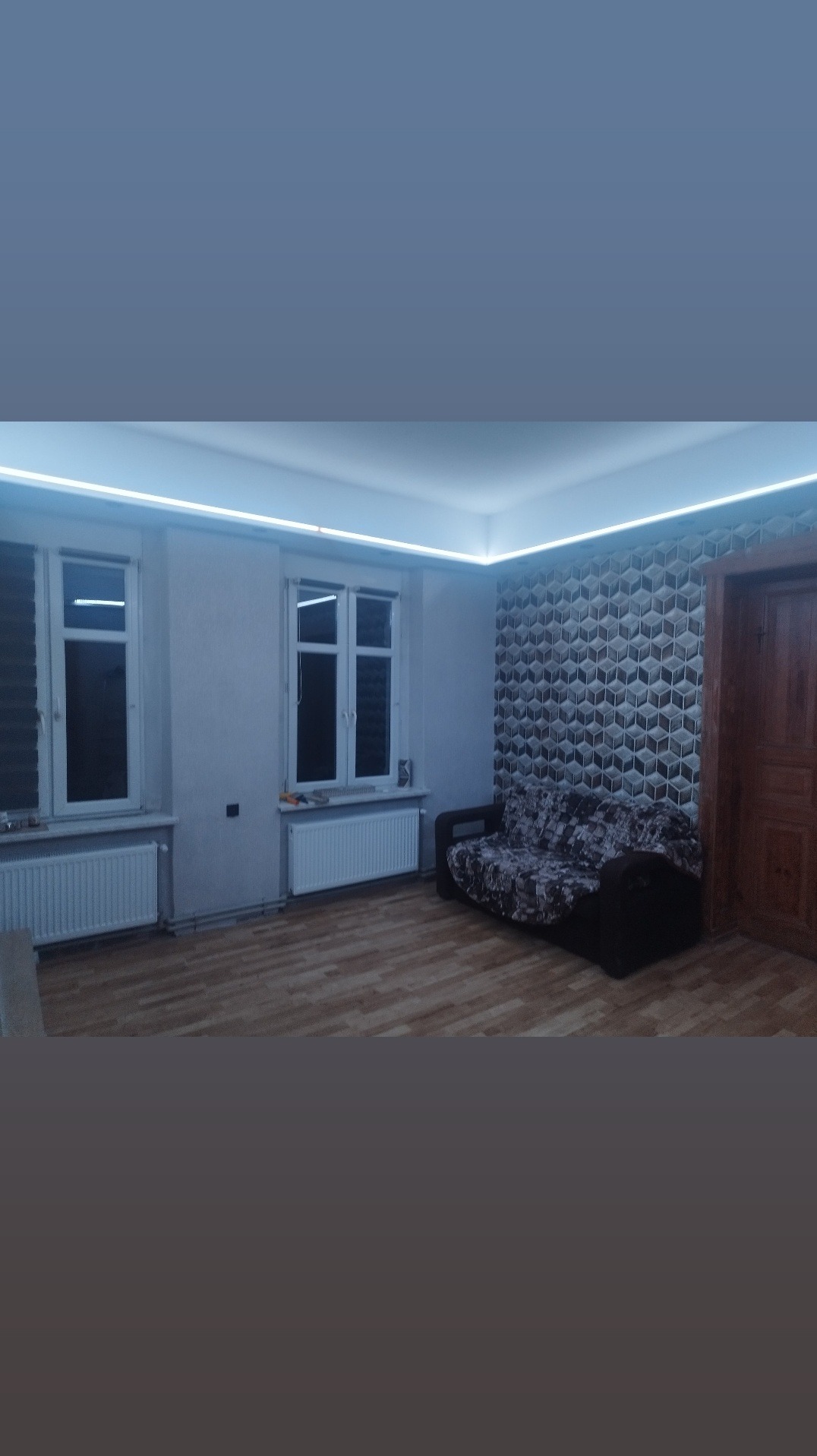 Pokój po remoncie z podświetleniem LED sufitu, wzorzystą tapetą 3D, drewnianą podłogą i grzejnikami pod oknami.
