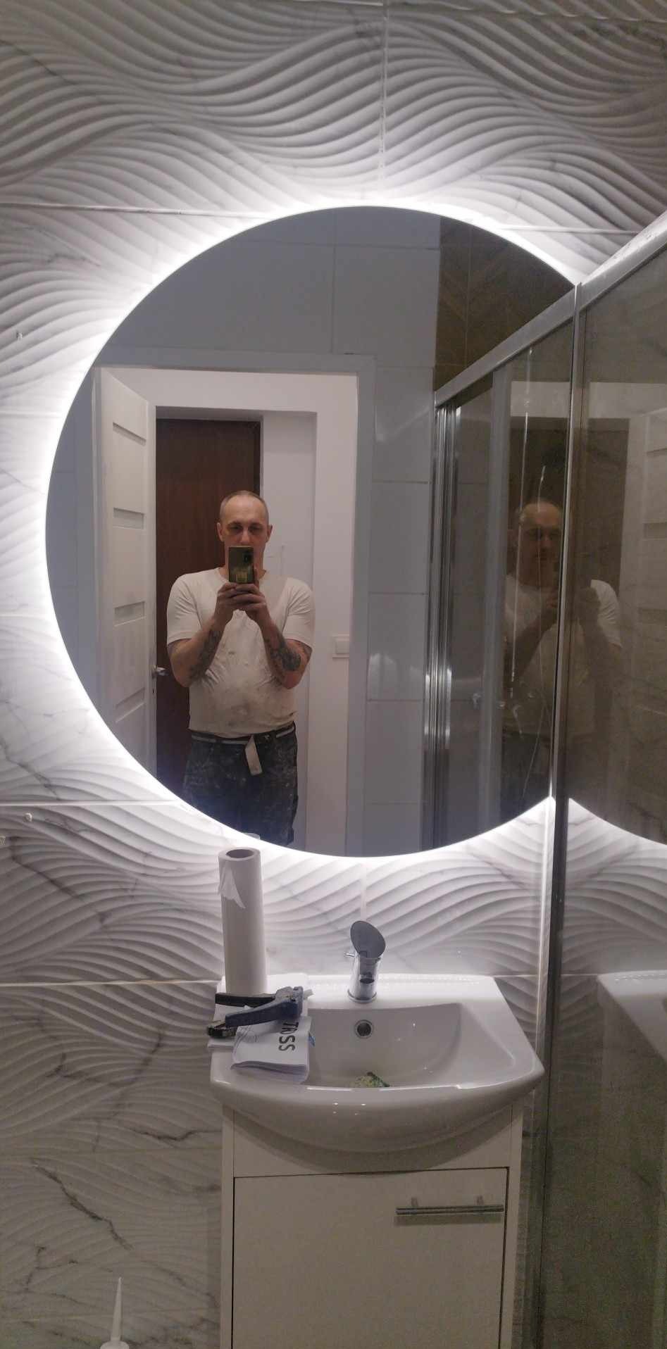 Okrągłe lustro z podświetleniem LED w wykończonej łazience z falistą, białą płytką na ścianie, widoczna umywalka z szafką i kabina prysznicowa, odbicie glazurnika robiącego zdjęcie.