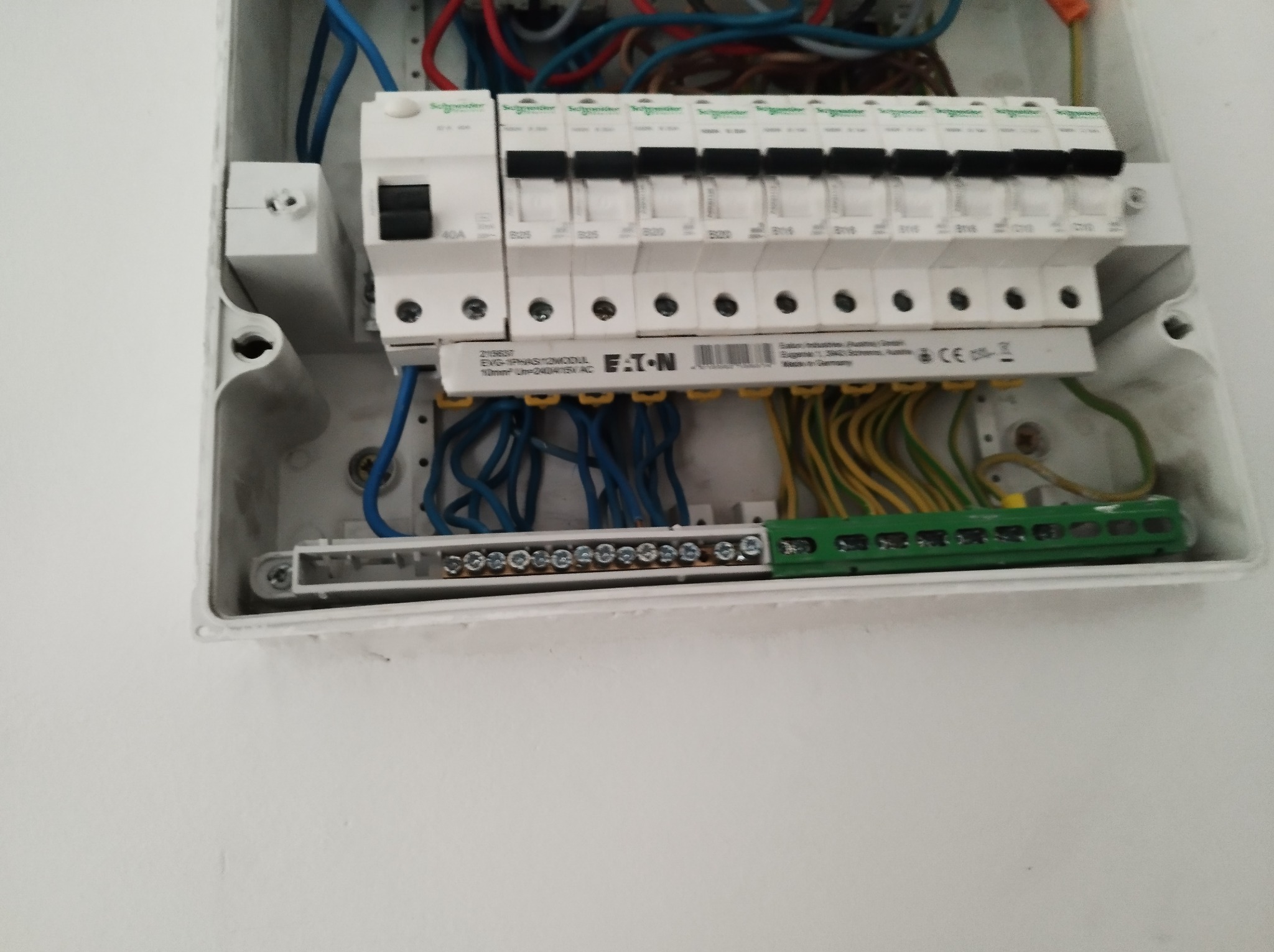 Otwarta skrzynka elektryczna z widocznymi bezpiecznikami, przewodami w kolorach niebieskim, czerwonym, żółto-zielonym, oraz listwą zaciskową z licznymi śrubami, zamontowana na białej ścianie.