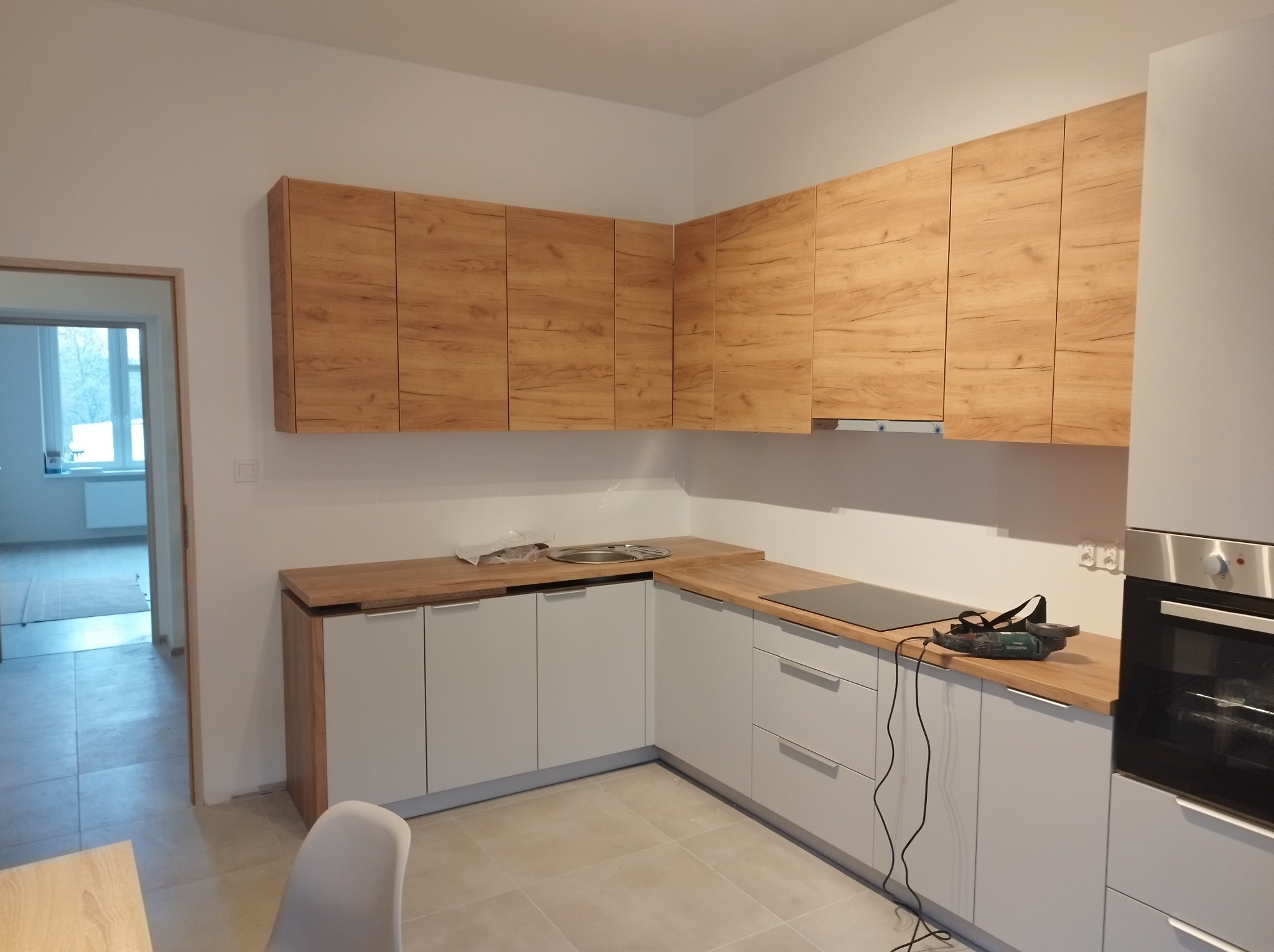 Nowoczesna kuchnia z szarymi szafkami dolnymi i drewnopodobnymi szafkami górnymi, widoczny blat z płytą indukcyjną i narzędzie na nim, jasna podłoga