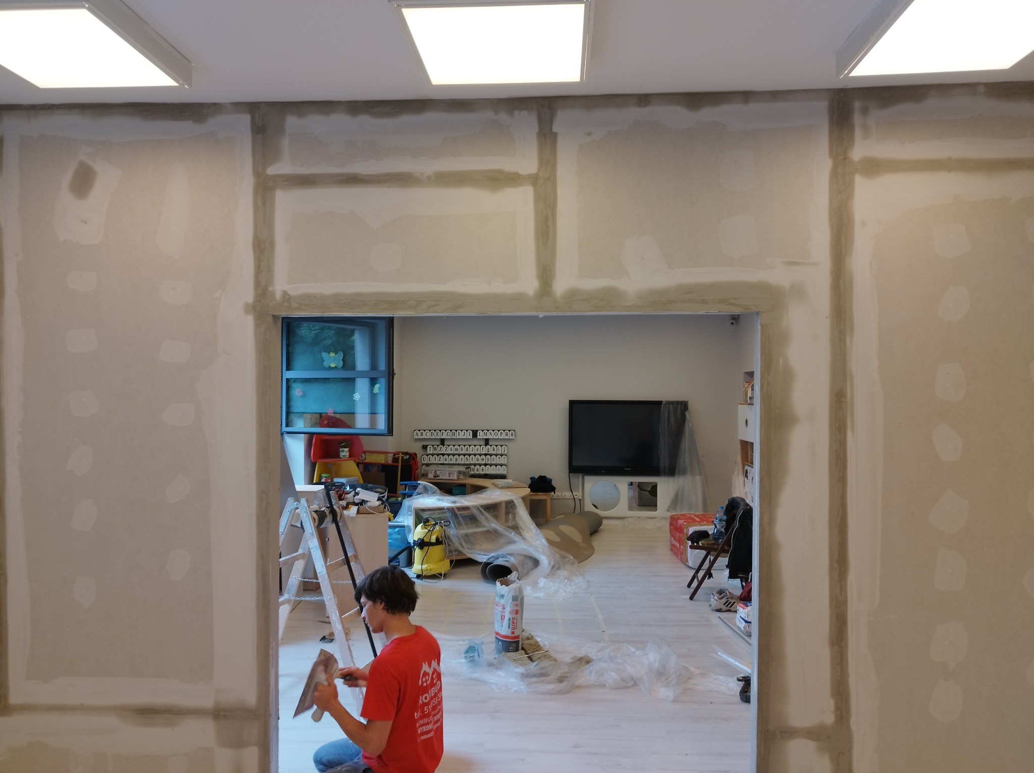 Prace wykończeniowe w pokoju: ściany z płyt kartonowo-gipsowych, częściowo pokryte masą szpachlową, widoczny pracownik z kielnią, przygotowujący powierzchnię do dalszych prac, wnętrze zabezpieczone...