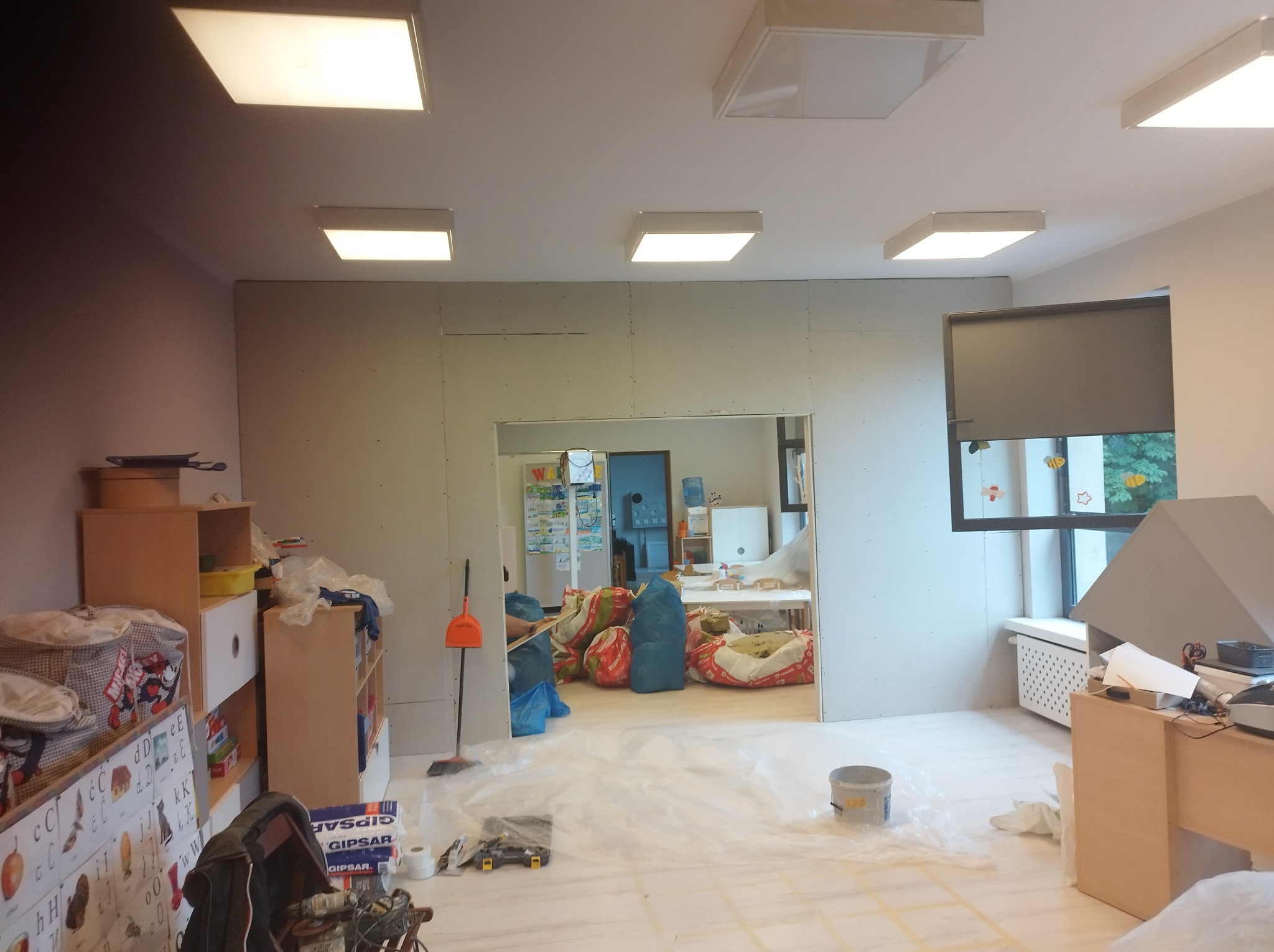 Montaż płyt karton gips na ścianie w trakcie remontu pokoju dziecięcego, widoczne narzędzia i worki z materiałem budowlanym, zabezpieczona folią podłoga.