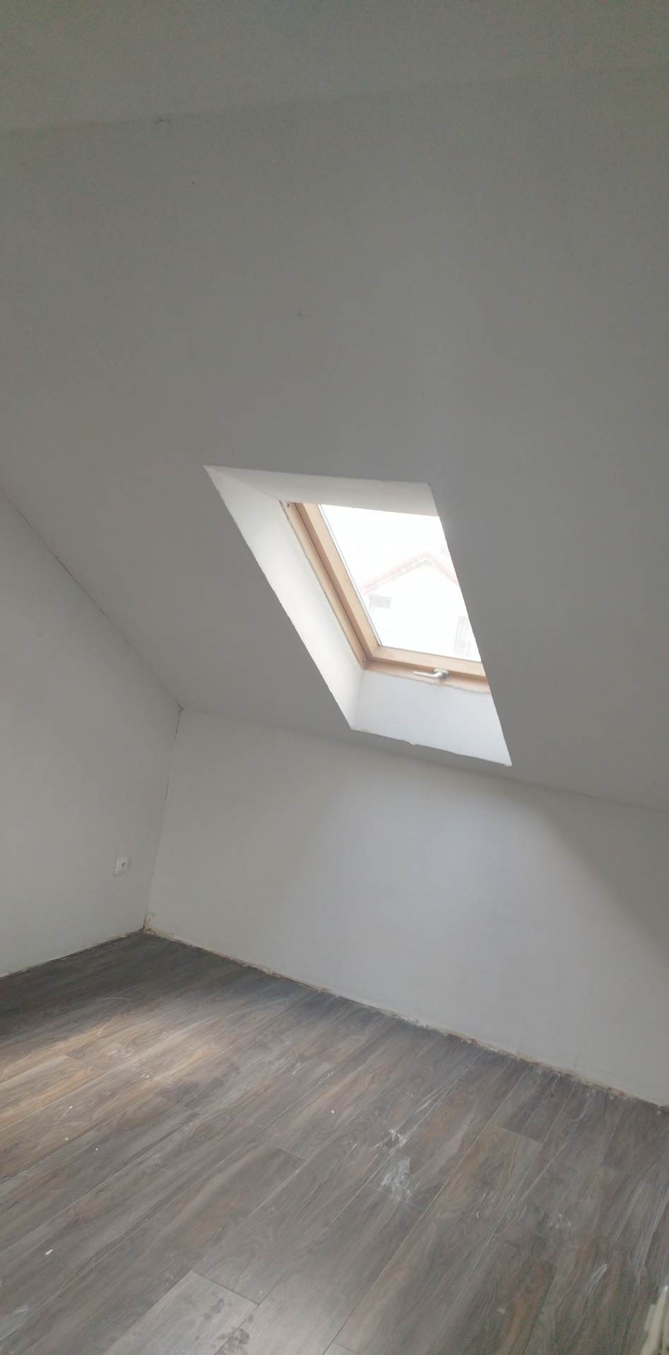 Poddasze po remoncie z drewnianymi panelami podłogowymi i oknem dachowym w białej ramie, ściany pomalowane na biało, widoczne ślady po remoncie przy podłodze.