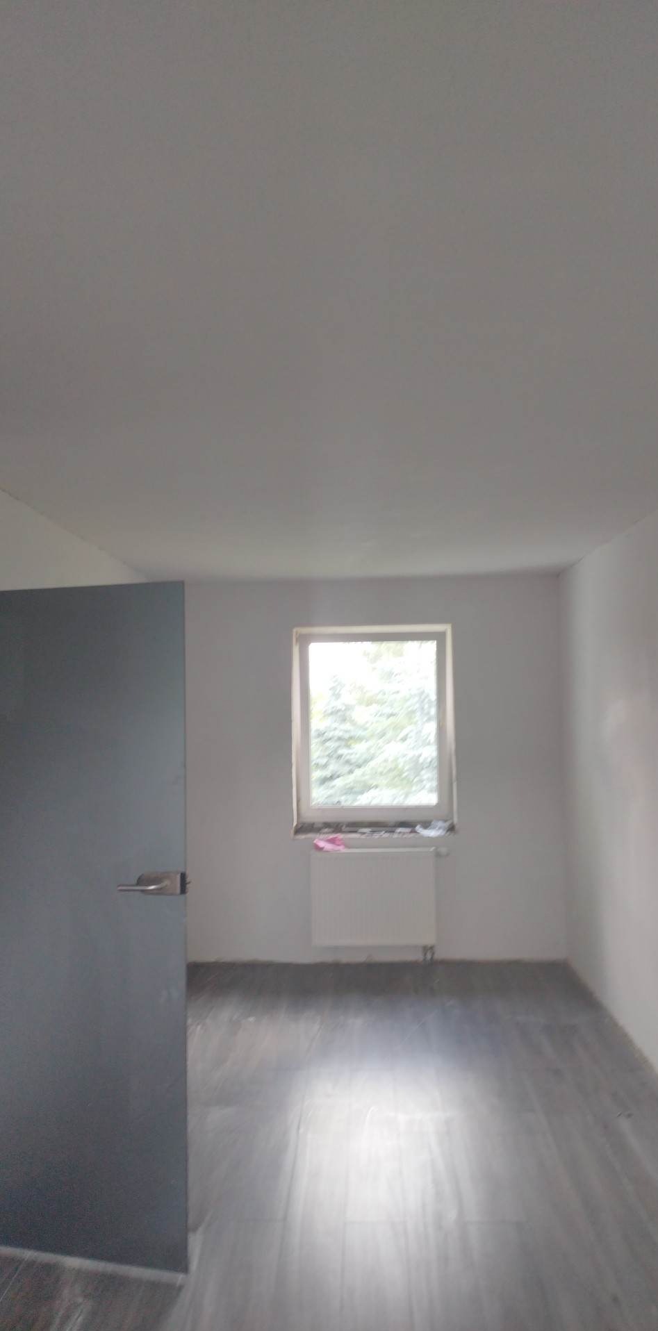 Pusty pokój po remoncie z szarą podłogą z paneli, białymi ścianami i grzejnikiem pod oknem, widok na częściowo otwarte drzwi.