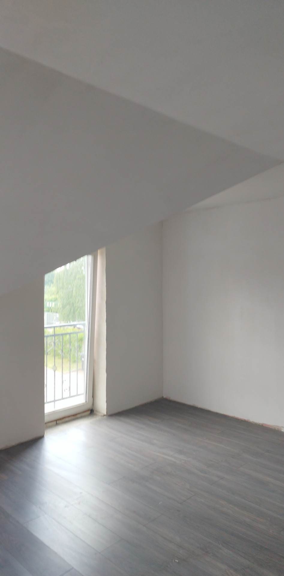 Poddasze po remoncie: świeżo położone ciemne panele podłogowe, białe ściany i sufit, okno balkonowe z metalową balustradą, widok na zieleń na zewnątrz.