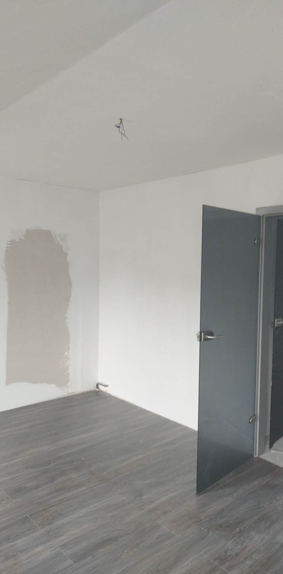 Pokój w trakcie remontu: szare panele podłogowe, ściana z fragmentem nałożonej szarej gładzi, biała ściana i sufit z wystającymi kablami, szklane drzwi.