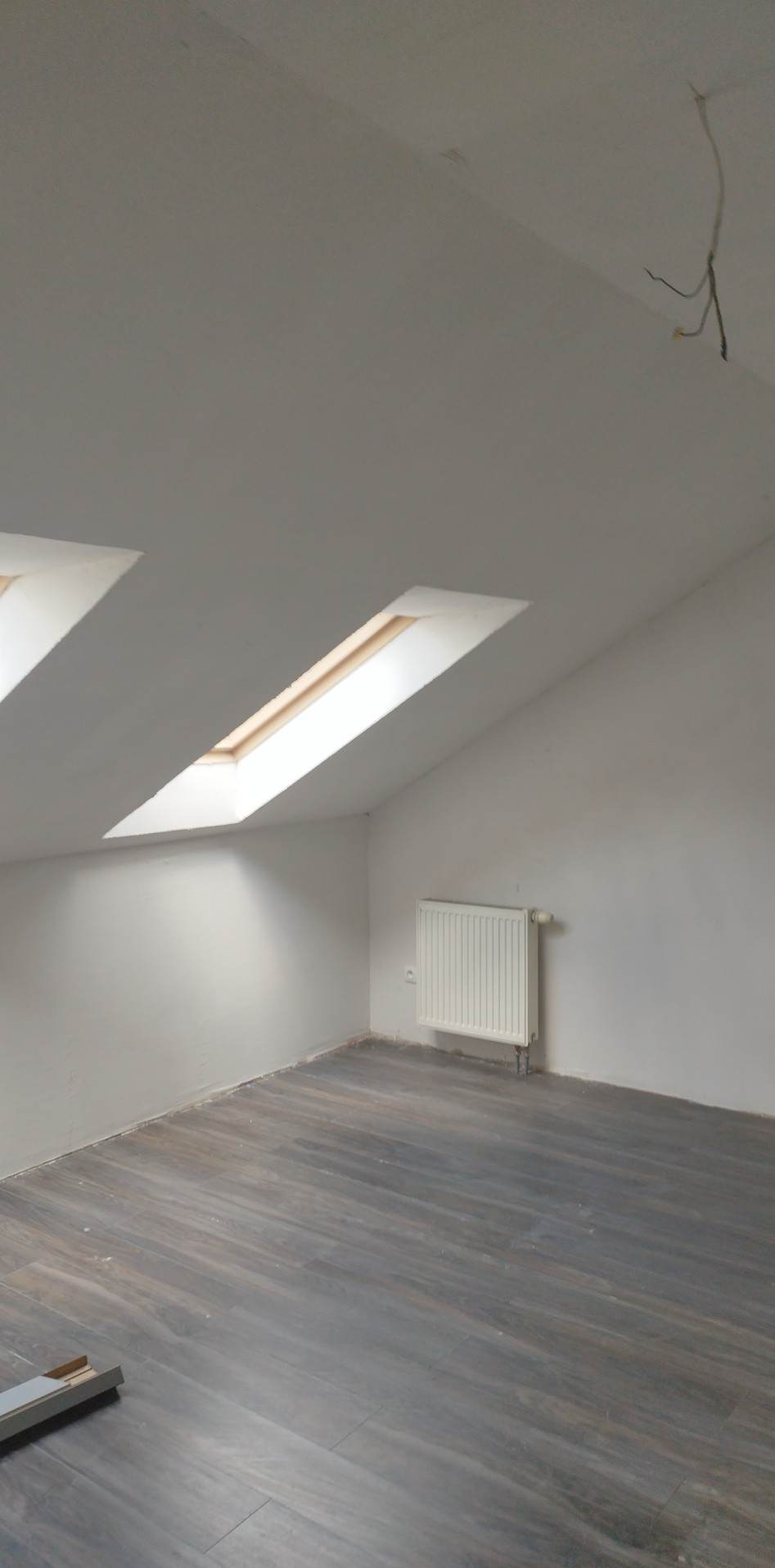 Pokój na poddaszu z dwoma oknami dachowymi, szarą podłogą imitującą drewno i białymi ścianami z grzejnikiem. Widoczne niedokończone wykończenie sufitu z wystającymi kablami.