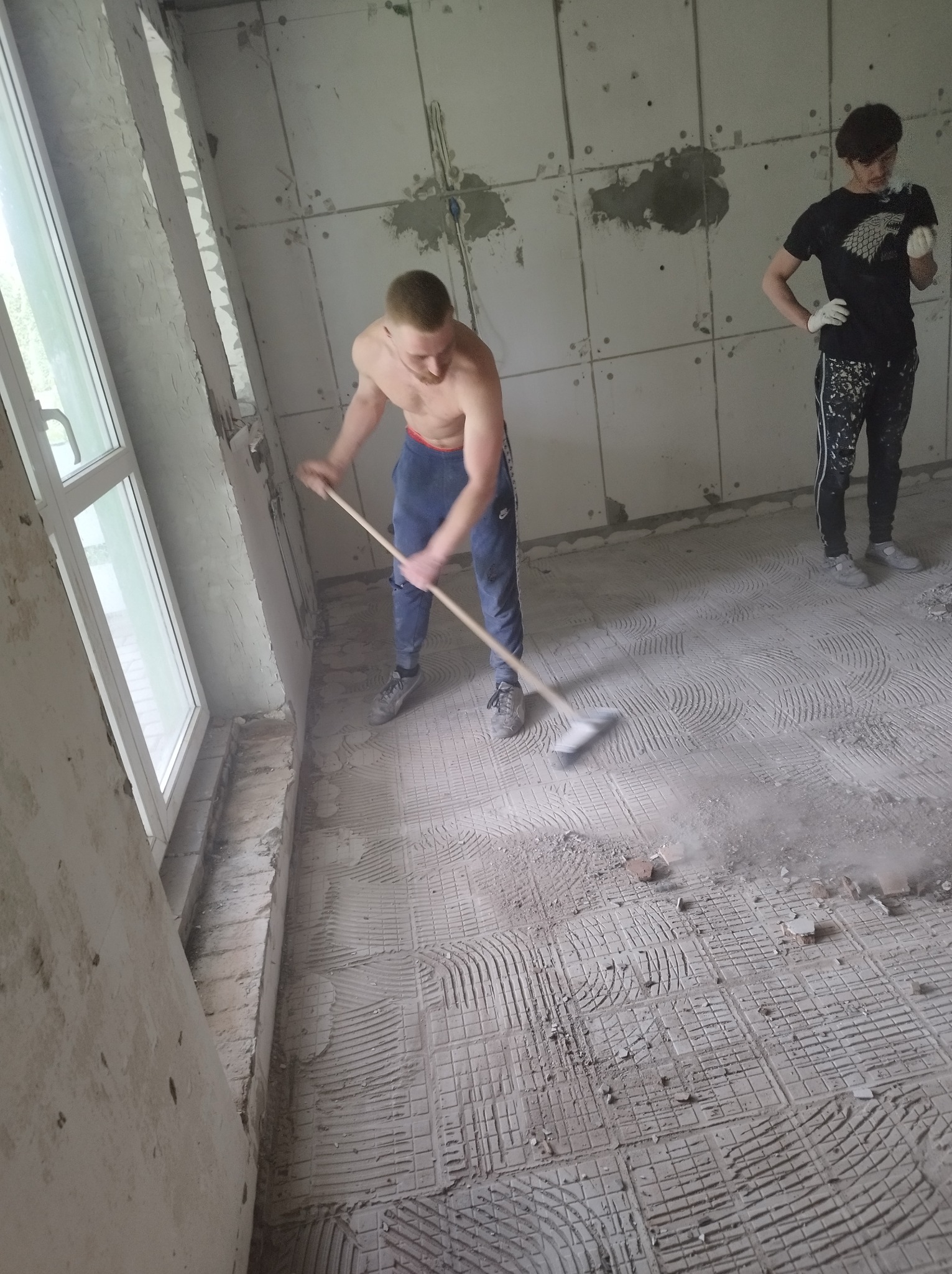 Mężczyzna zamiatający podłogę po skuciu płytek w remontowanym pomieszczeniu z oknem i ścianami wyłożonymi płytami karton gips.