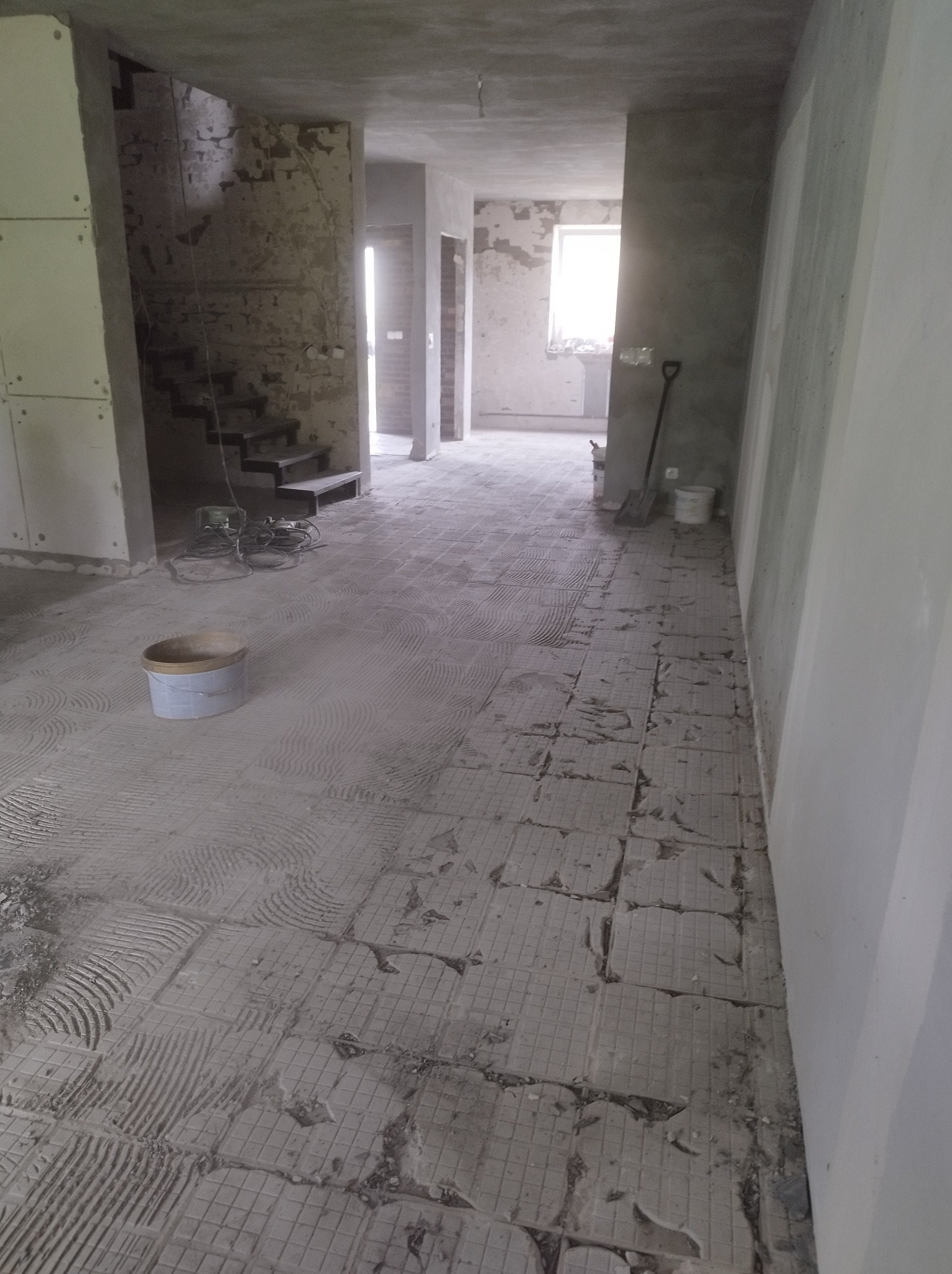Wnętrze w trakcie prac remontowych: zerwane płytki podłogowe z widocznymi śladami kleju, wiadro z zaprawą, narzędzia, surowe ściany częściowo pokryte płytami gipsowo-kartonowymi, schody w tle.