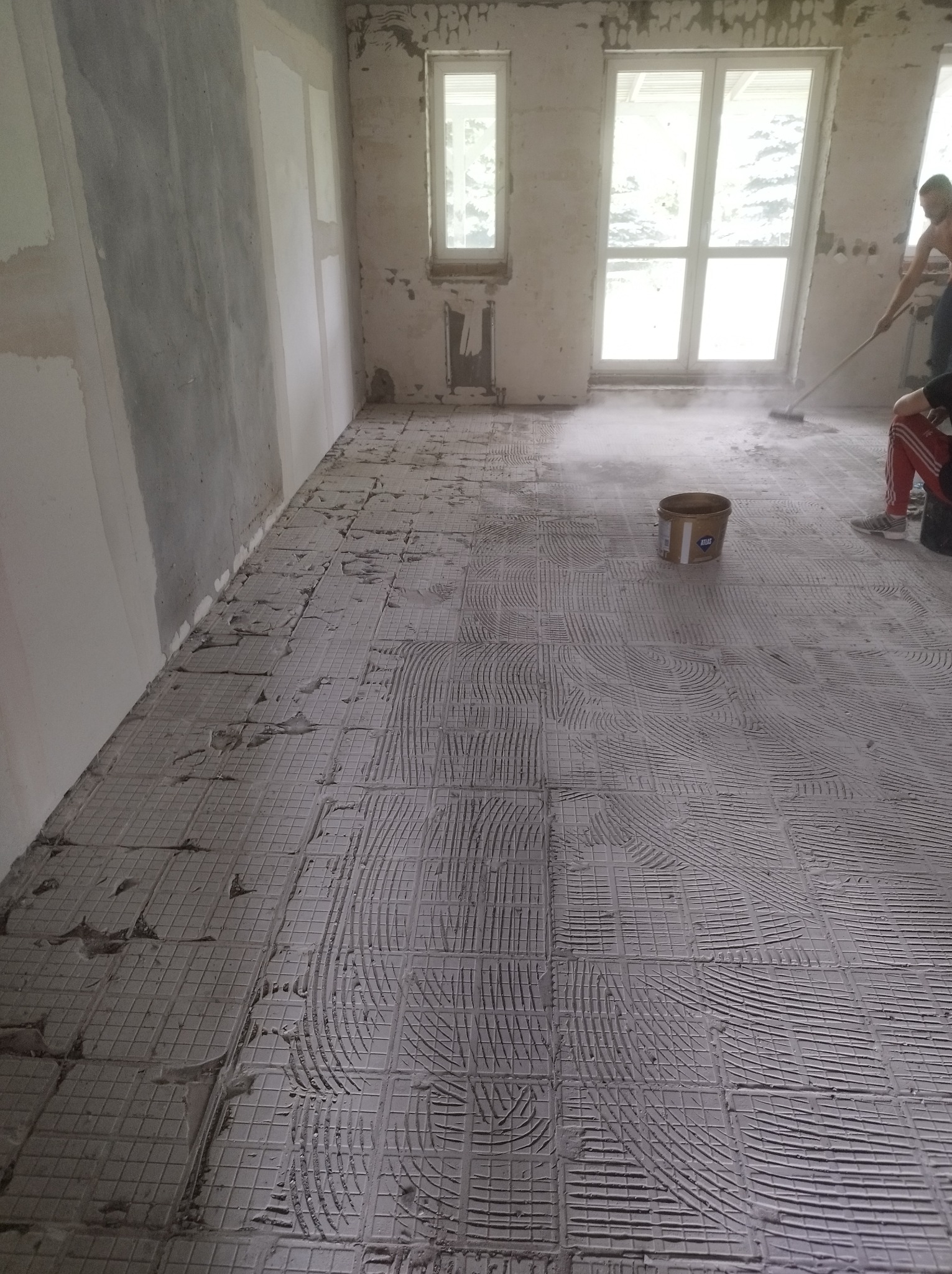 Przygotowanie podłoża pod płytki: warstwa kleju rozprowadzona na podłodze z widocznymi śladami pacy zębowej, wiadro z klejem Atlas w tle, fragment ściany z widocznymi warstwami gładzi.