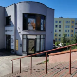 Zewnętrzny widok budynku usługowego z reklamą w oknach przedstawiającą zabieg na twarzy, widoczne schody wejściowe z barierką.