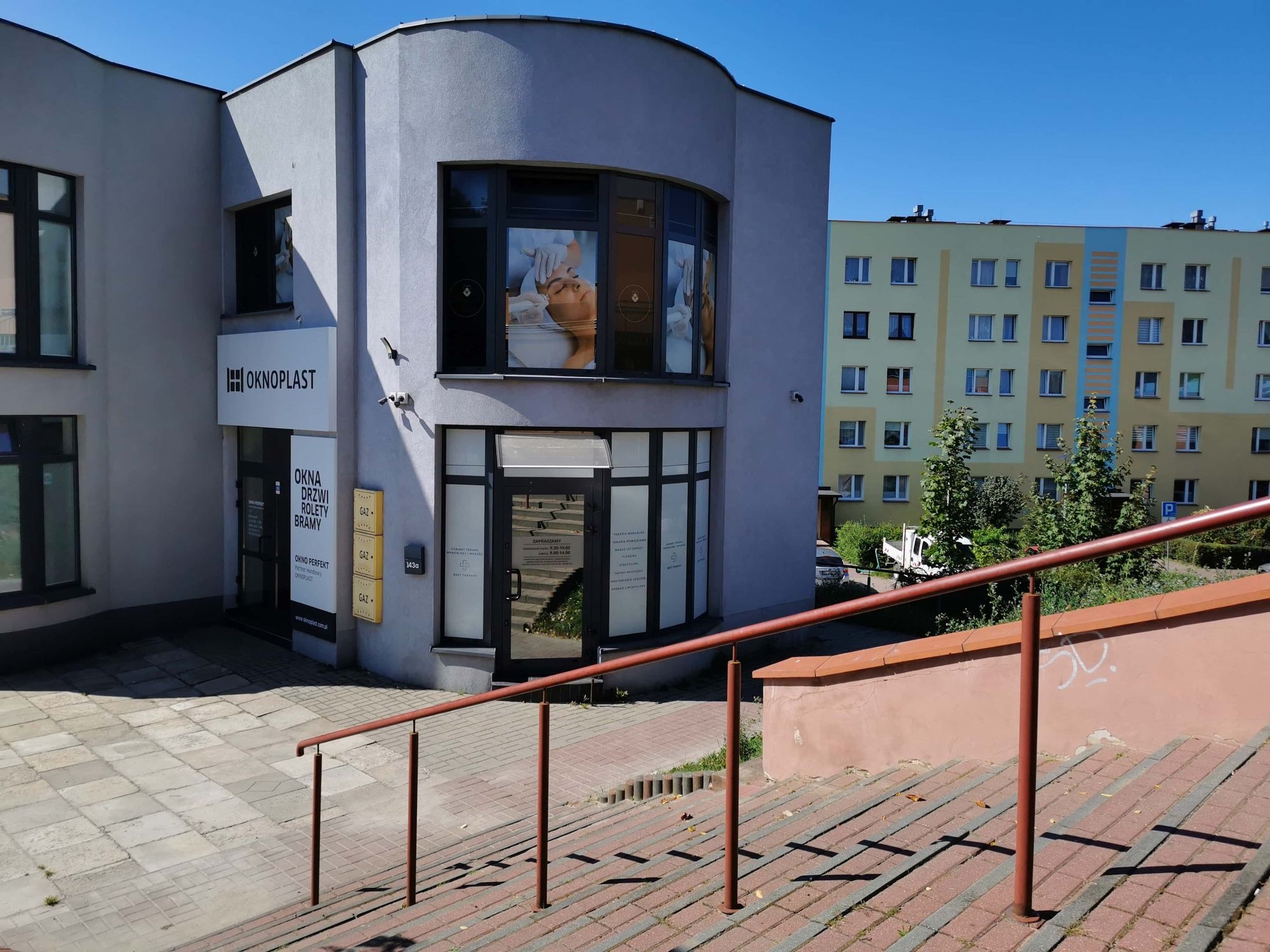 Zewnętrzny widok budynku usługowego z reklamą w oknach przedstawiającą zabieg na twarzy, widoczne schody wejściowe z barierką.