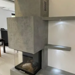 Nowoczesny kominek z szarego betonu architektonicznego, z wkładem kominkowym obudowanym szkłem, w minimalistycznym wnętrzu.