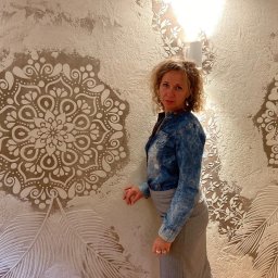 Virya- Wnętrza Liliana Strzelecka - Kobieta w jeansowej koszuli pozuje na tle ściany z dekoracyjną, strukturalną gładzią w odcieniach bieli i beżu, z motywami mandali i liści.