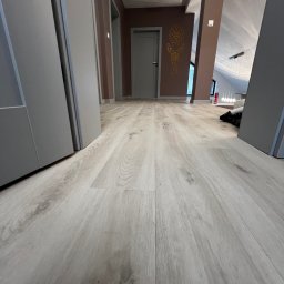 Virya- Wnętrza Liliana Strzelecka - Jasne panele podłogowe w korytarzu z szarą zabudową, perspektywa z niskiego kąta, widoczne drzwi i dekoracja na ścianie w głębi.