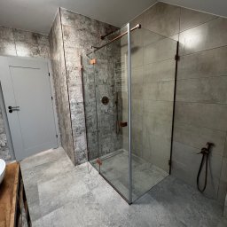 Virya- Wnętrza Liliana Strzelecka - Nowoczesna łazienka z kabiną prysznicową typu walk-in, szare płytki imitujące beton, armatura w kolorze miedzi i drewniana szafka pod umywalką. Minimalistyczny design.