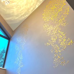 Virya- Wnętrza Liliana Strzelecka - Dekoracyjna ściana z ornamentami roślinnymi w odcieniach złota, oświetlona nowoczesną lampą. Widok z dołu, z fragmentem okna po lewej stronie.
