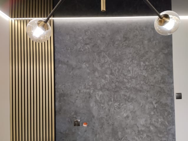 Ściana z dekoracyjną szarą strukturą, oświetlona listwą LED i lampą z kulistymi kloszami, obok ściana wykończona pionowymi panelami z jasnego drewna.