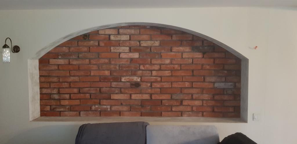 Wnęka z cegły klinkierowej w łukowatym kształcie, otoczona szarą ramą, z widoczną lampką ścienną po lewej stronie i fragmentem sofy na dole.