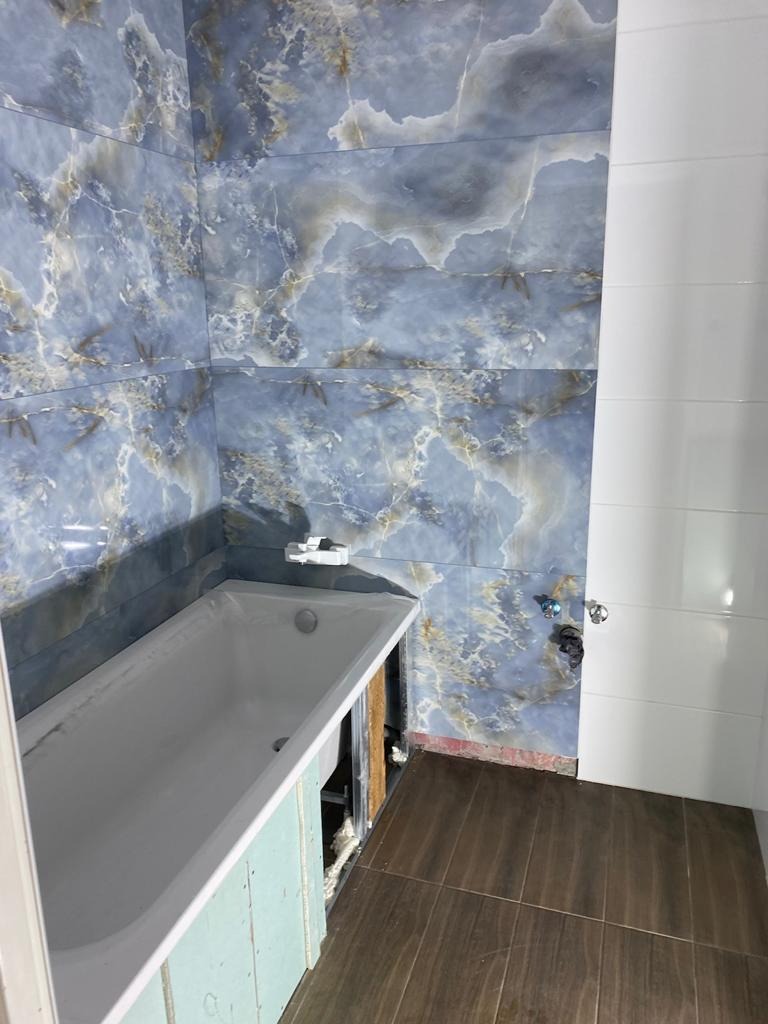Niedokończona łazienka z wanną obudowaną płytami, marmurowe płytki ścienne w odcieniach niebieskiego i beżu, oraz brązowe płytki podłogowe imitujące drewno.
