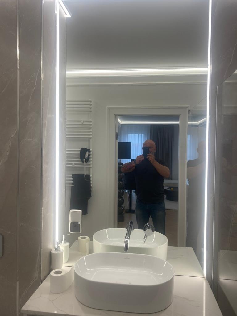 Nowoczesna łazienka z podwójną umywalką nablatową, marmurowymi płytkami ściennymi i lustrem z oświetleniem LED, widoczne akcesoria łazienkowe i grzejnik drabinkowy w tle, odbicie osoby robiącej...