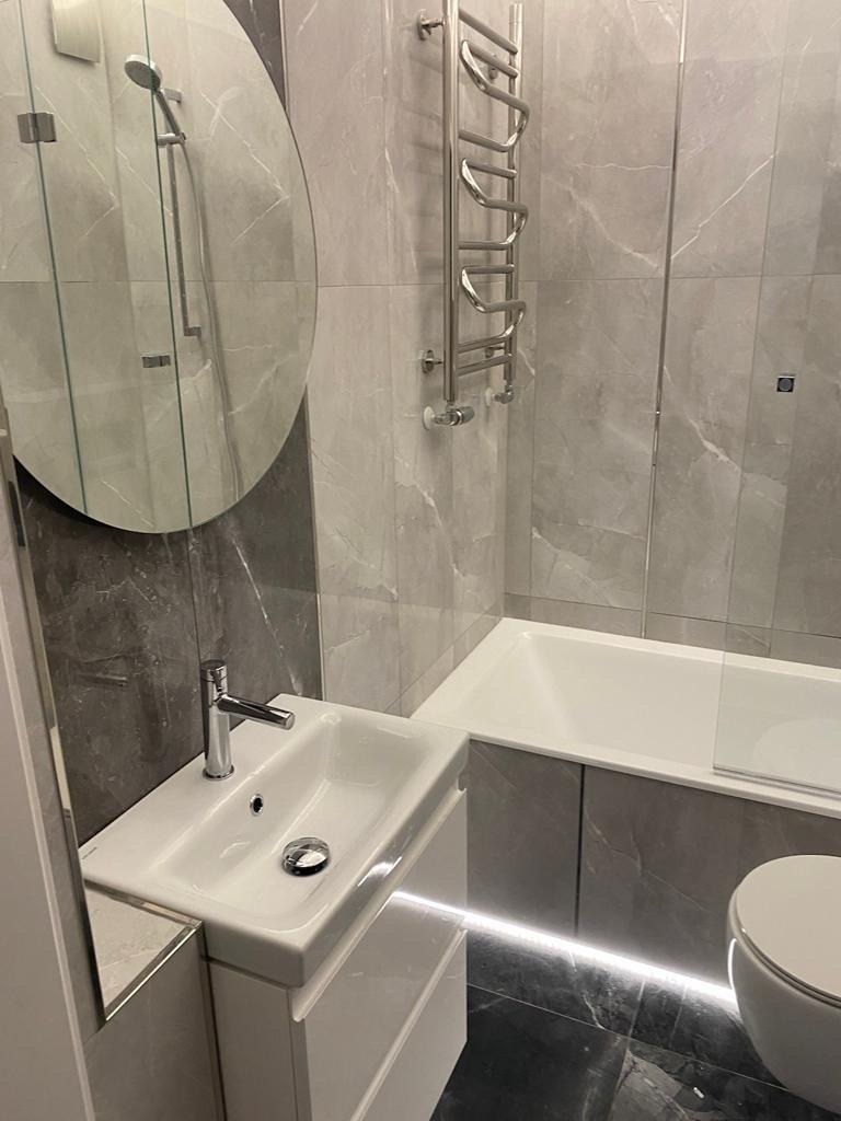 Nowoczesna łazienka z marmurowymi płytkami, podświetlaną wanną, owalnym lustrem i małą umywalką z szafką