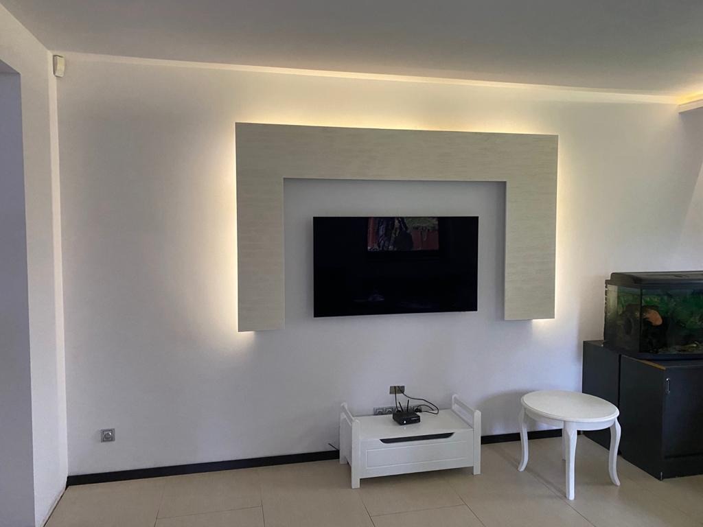 Salon z telewizorem wkomponowanym w podświetlaną ramę z płyty, poniżej biała szafka RTV i stolik, w tle akwarium.