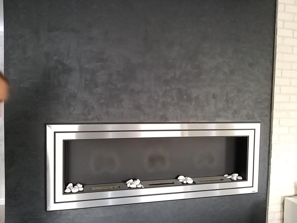 Ściana z efektem dekoracyjnym w odcieniach szarości, z wbudowanym biokominkiem obłożonym białymi kamieniami; obok fragment ściany z białej cegły.