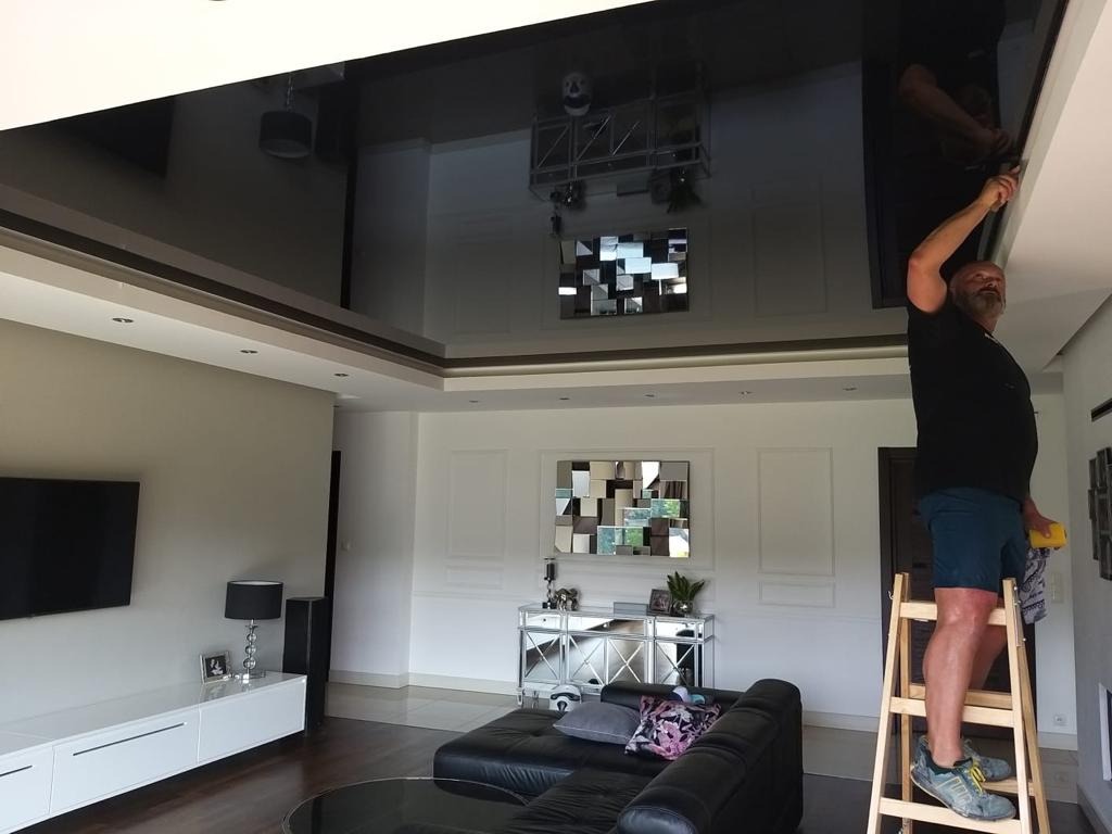 Mężczyzna na drewnianej drabinie czyści błyszczący, czarny sufit w luksusowym salonie, widoczne odbicie wyposażenia i oświetlenia.