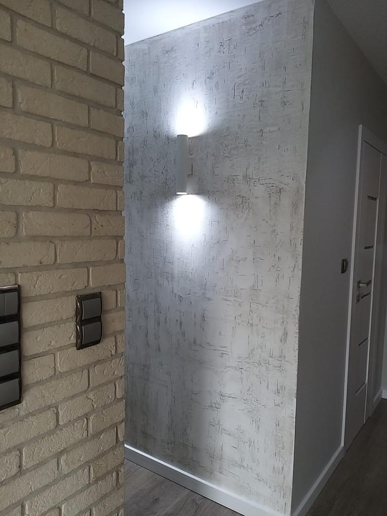 Korytarz z dekoracyjną, szarą ścianą o fakturze betonu, oświetloną przez dwa pionowe, cylindryczne kinkiety. Po lewej widoczna ściana z białej cegły z czarnymi włącznikami.