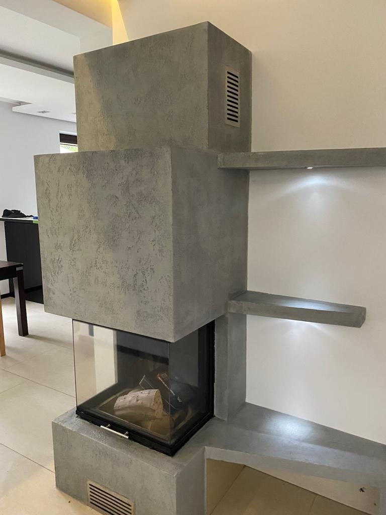 Nowoczesny kominek z szarego betonu architektonicznego, z wkładem kominkowym obudowanym szkłem, w minimalistycznym wnętrzu.