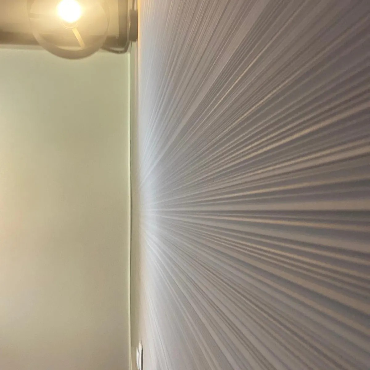 Ściana w pokoju z tapetą w geometryczny wzór rozchodzących się promieni, oświetlona lampą sufitową z żarówką.