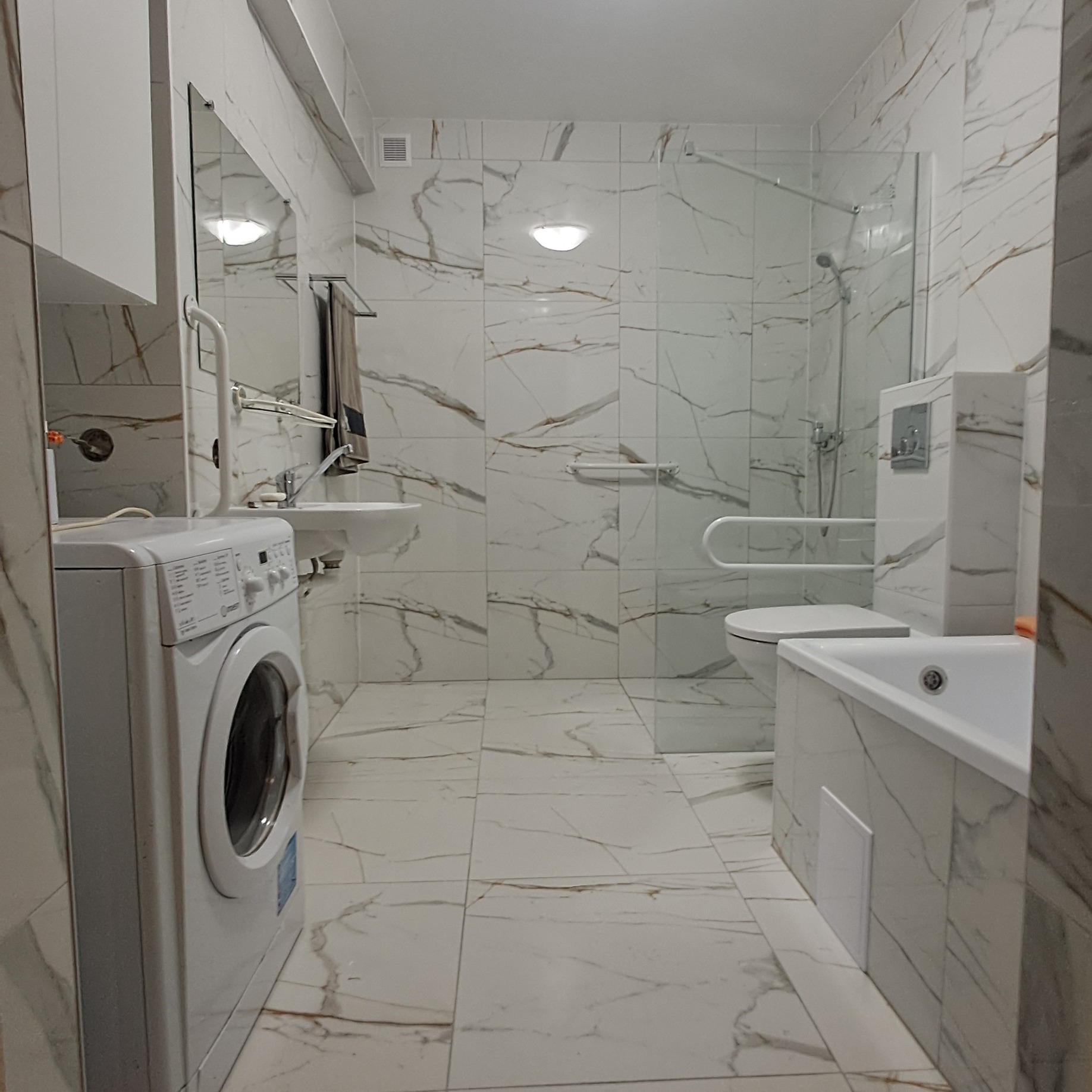 Wykończona łazienka w jasnych płytkach imitujących marmur, z umywalką, pralką, toaletą z uchwytami dla niepełnosprawnych i kabiną prysznicową.