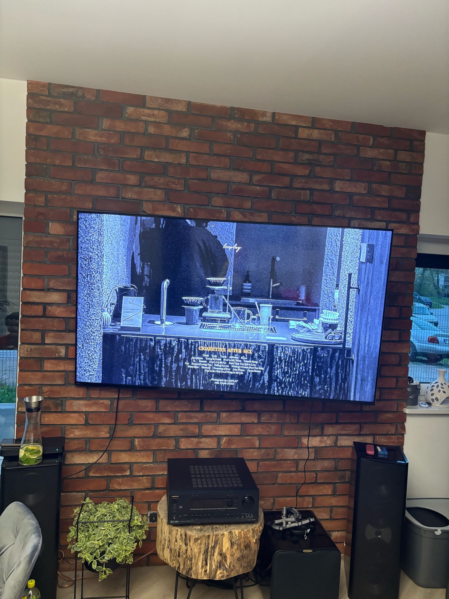 Telewizor wiszący na ścianie z czerwonej cegły, wyświetlający czarno-biały obraz wnętrza z blatem i akcesoriami, poniżej sprzęt audio na drewnianym stoliku, obok głośniki i doniczka z rośliną.