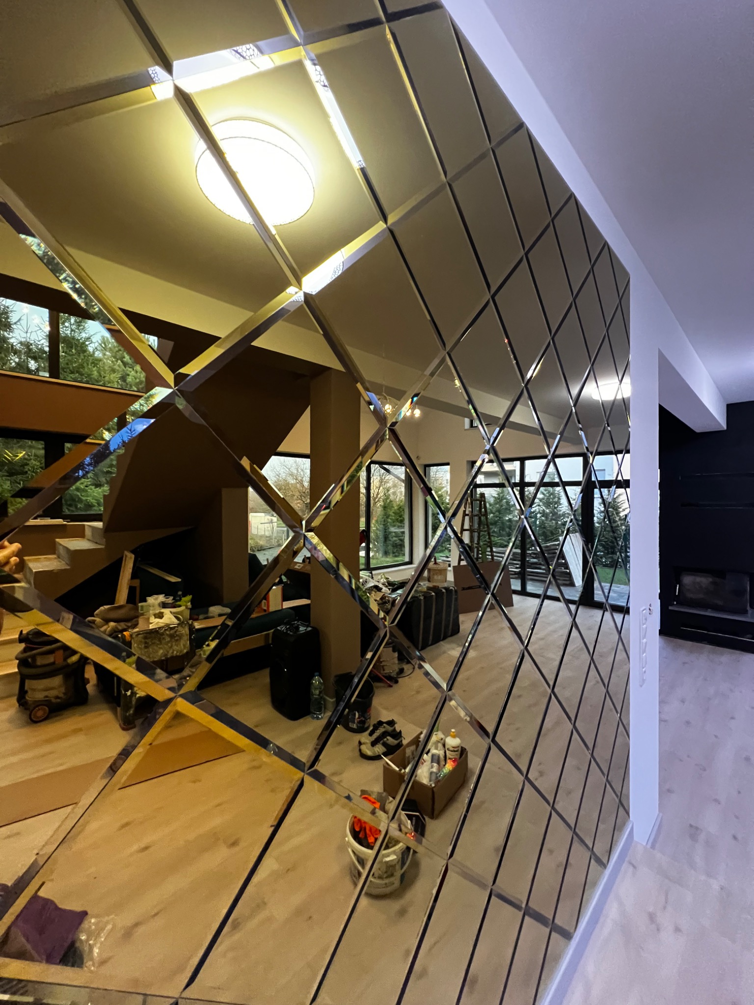 Ściana z dekoracyjnych, lustrzanych płytek w romby, odbijająca wnętrze domu z drewnianymi schodami i oknami. Widoczne narzędzia i materiały budowlane.