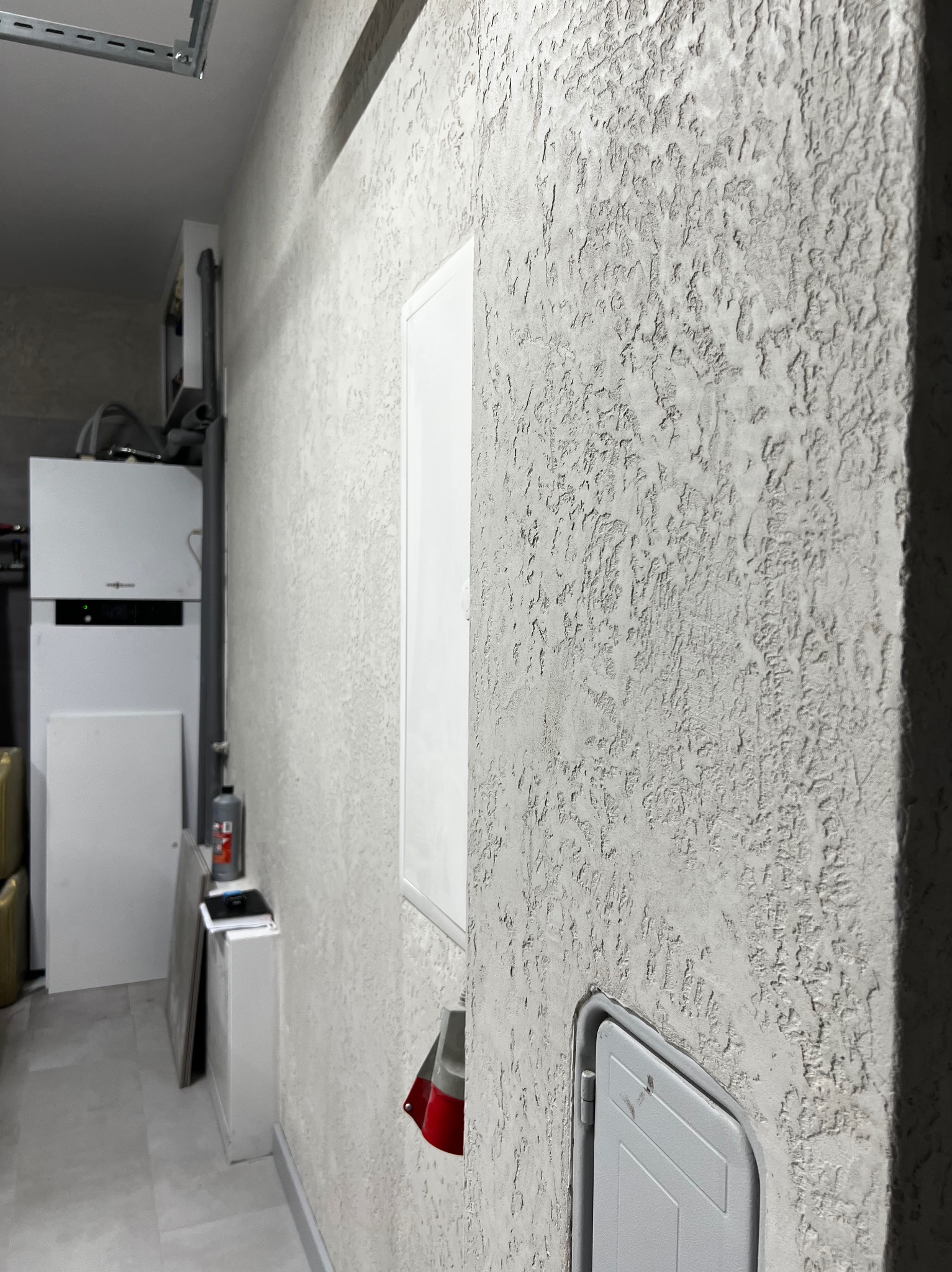 Ściana z fakturą baranka w odcieniach szarości, z widoczną białą skrzynką elektryczną i fragmentem instalacji grzewczej w tle. Minimalistyczny design.