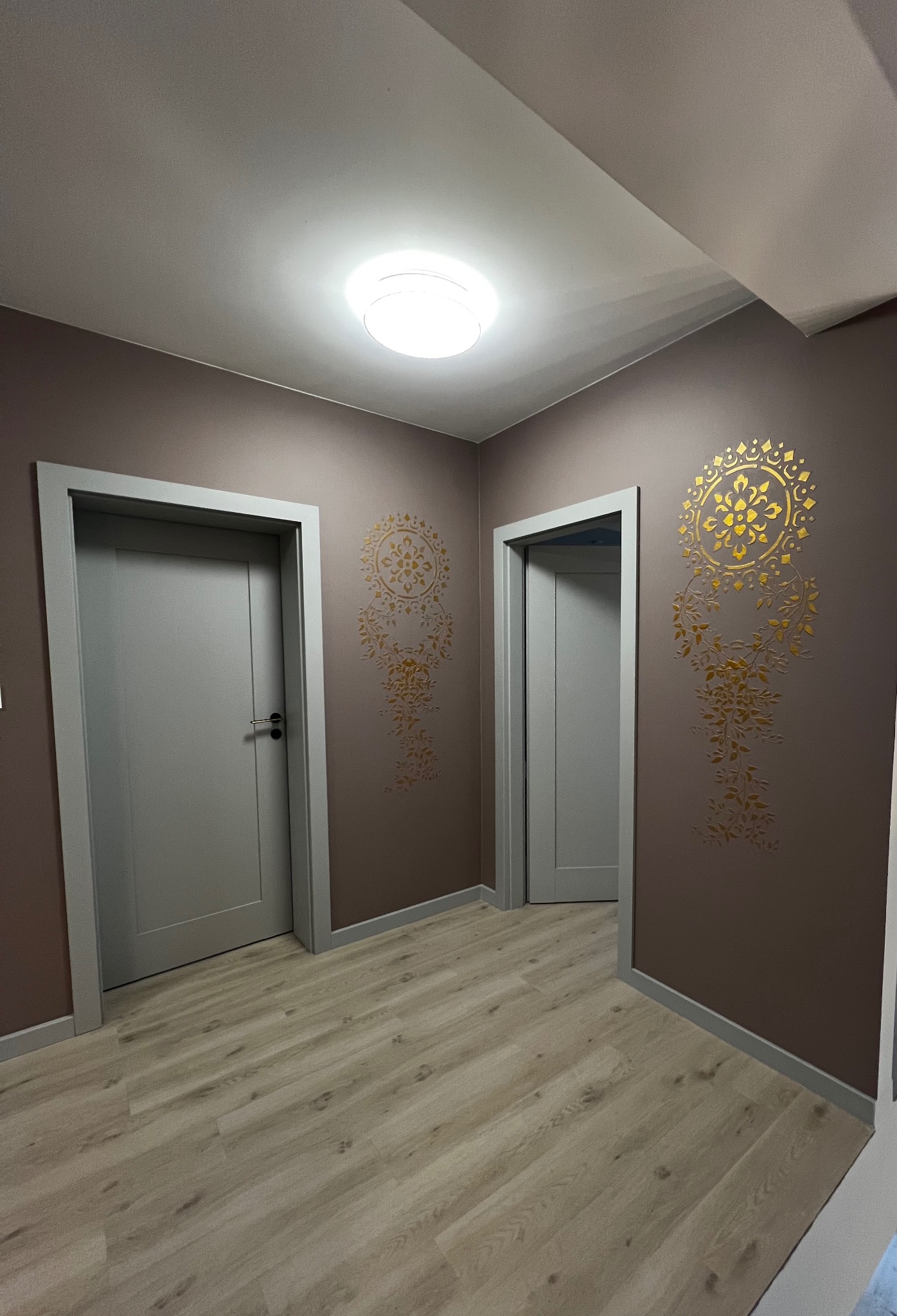Korytarz po remoncie: brązowe ściany zdobione złotym ornamentem, jasna podłoga z paneli, dwoje szarych drzwi z ościeżnicami. Nowoczesne oświetlenie sufitowe.