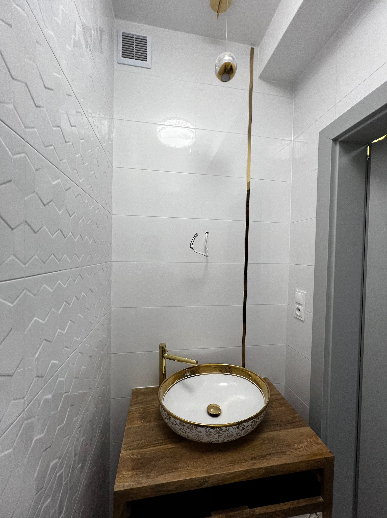 Elegancka łazienka po remoncie: białe płytki z geometrycznym wzorem, złota armatura i dekoracyjna umywalka na drewnianym blacie. Widoczne detale wykończenia.
