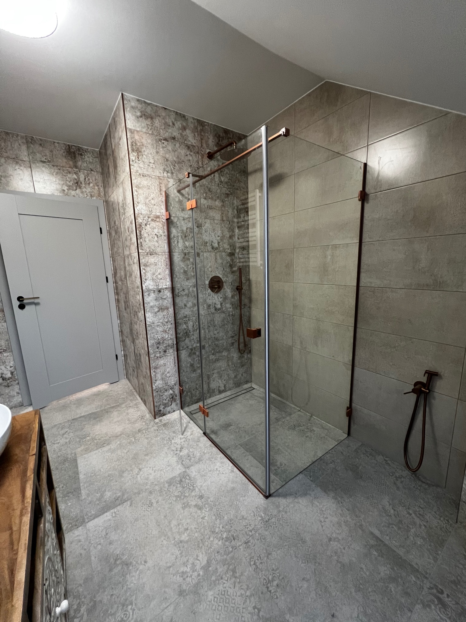 Nowoczesna łazienka z kabiną prysznicową typu walk-in, szare płytki imitujące beton, armatura w kolorze miedzi i drewniana szafka pod umywalką. Minimalistyczny design.