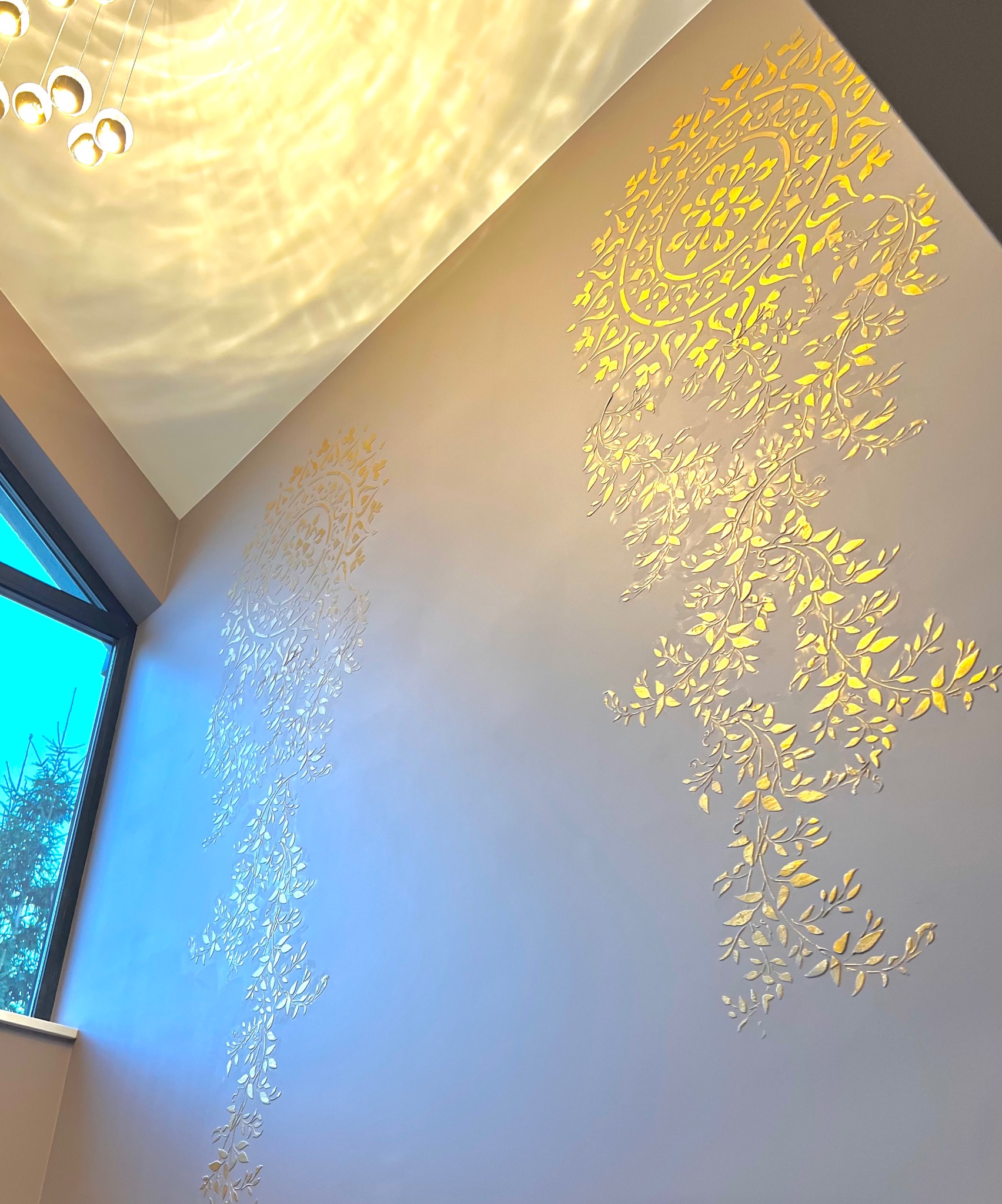 Dekoracyjna ściana z ornamentami roślinnymi w odcieniach złota, oświetlona nowoczesną lampą. Widok z dołu, z fragmentem okna po lewej stronie.