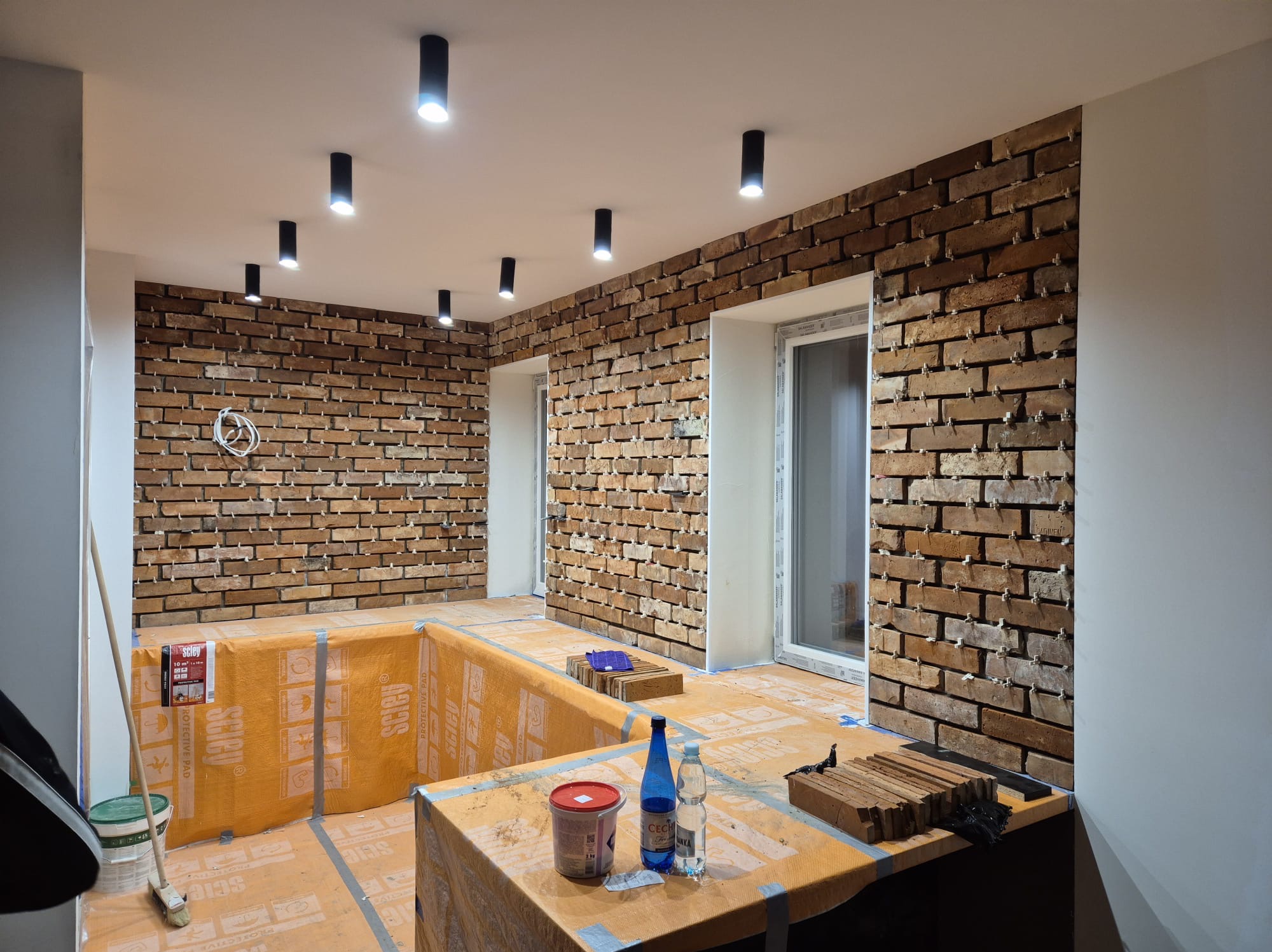 Ściana z cegły klinkierowej w trakcie montażu, widoczne krzyżyki dystansowe, zabezpieczona podłoga, oświetlenie punktowe sufitowe. Prace wykończeniowe.
