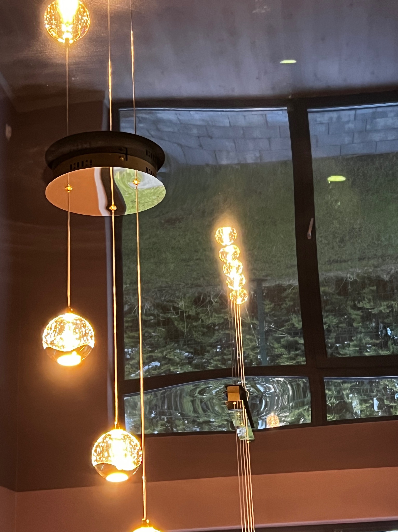 Nowoczesna lampa wisząca z kulistymi kloszami, odbijająca światło w oknie. Widok na zieleń za oknem. Ciepłe oświetlenie wnętrza.