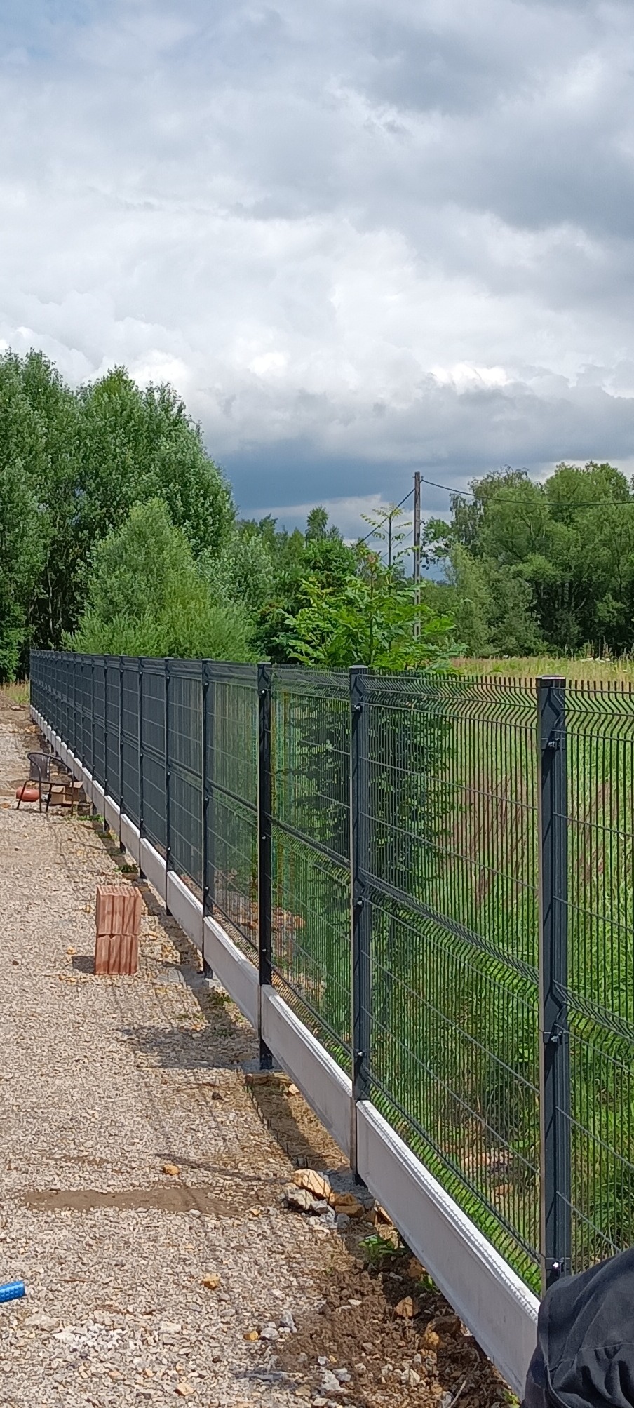 Długi, ciemnoszary system ogrodzeniowy z siatki panelowej na betonowej podmurówce, widok z boku na żwirowej ścieżce, z zielenią w tle i pochmurnym niebem.