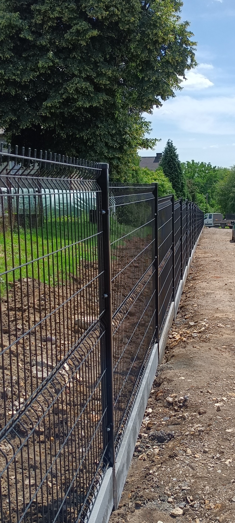 Nowo zamontowane, czarne ogrodzenie panelowe z betonową podmurówką na nierównym terenie z widoczną ziemią i kamieniami. W tle zieleń drzew i budynki.