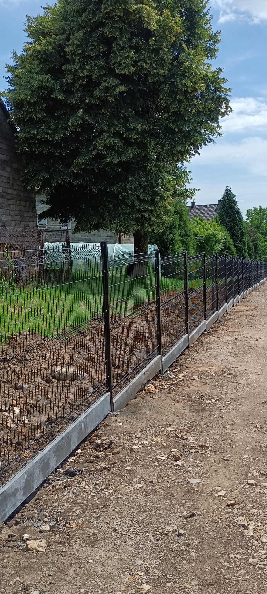 Czarny ogrodzenie panelowe z podmurówką betonową, widok z zewnątrz posesji, w tle drzewa i dom.