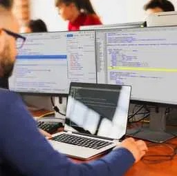 Programista z brodą pracuje przy biurku z dwoma monitorami i laptopem. Na ekranach widoczny kod źródłowy. Widoczny fragment klawiatury i myszki.