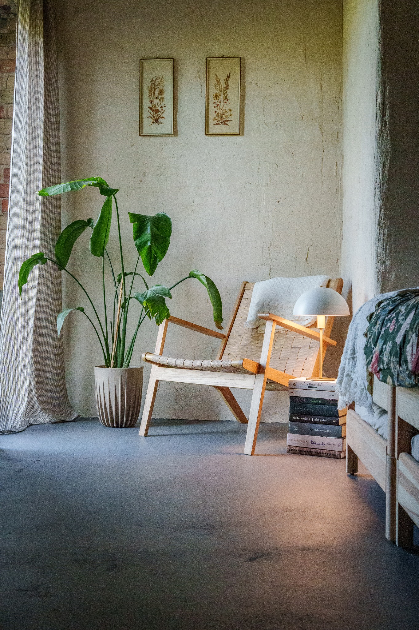 Jasne wnętrze z drewnianym fotelem i rośliną w doniczce. Na ścianie wiszą grafiki, a obok fotela leżą książki i stoi lampka. Przytulny klimat.